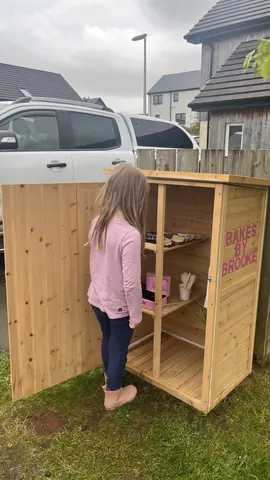 A girl is standing in front of a wooden box with a sign that says "Bakes by Brooke."