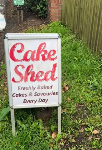 A sign for a cake shed is on a grassy area.