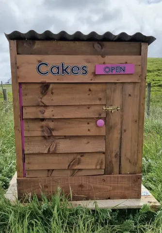 A wooden structure with a pink sign that says "Cakes Open" on it.