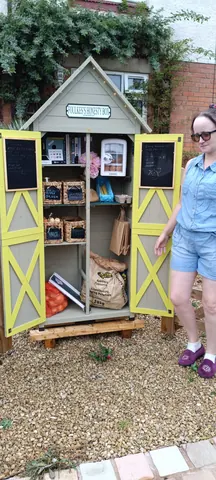 A woman standing in front of a small wooden structure with a bag of groceries.