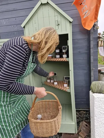 A woman is holding a basket and looking at a shelf with jars on it.