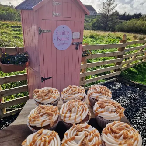 A pink shed with a sign that says "Smitty's Bakes" and a bunch of cupcakes in front of it.