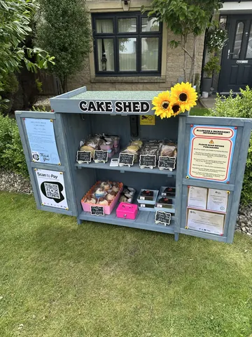 A small cake shop with a sign that says "Cake Shed" and a sunflower on it.