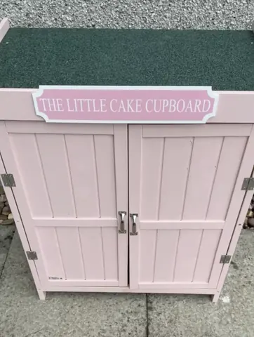 A pink cupboard with a sign on it that says "The Little Cake Cupboard".