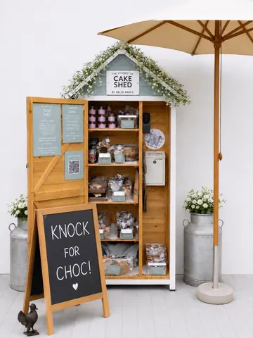 A small wooden shed with a sign that says "knock for choc!" and a cake display inside.